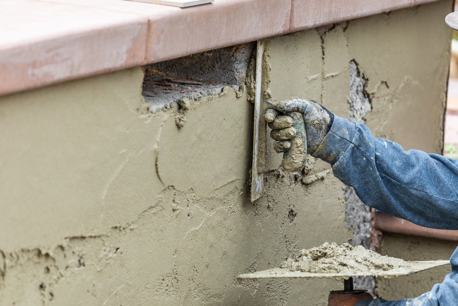 The image shows a person applying wet cement or stucco to a vertical surface with a construction trowel. The person is wearing a long sleeve shirt and protective gloves, which suggests they are adhering to safety practices typical for construction work. The surface appears to be an outdoor wall, and the person is in the process of smoothing out the mixture to either repair or create a smooth finish on the wall. The work seems to be meticulous, requiring skill to ensure an even and smooth application.