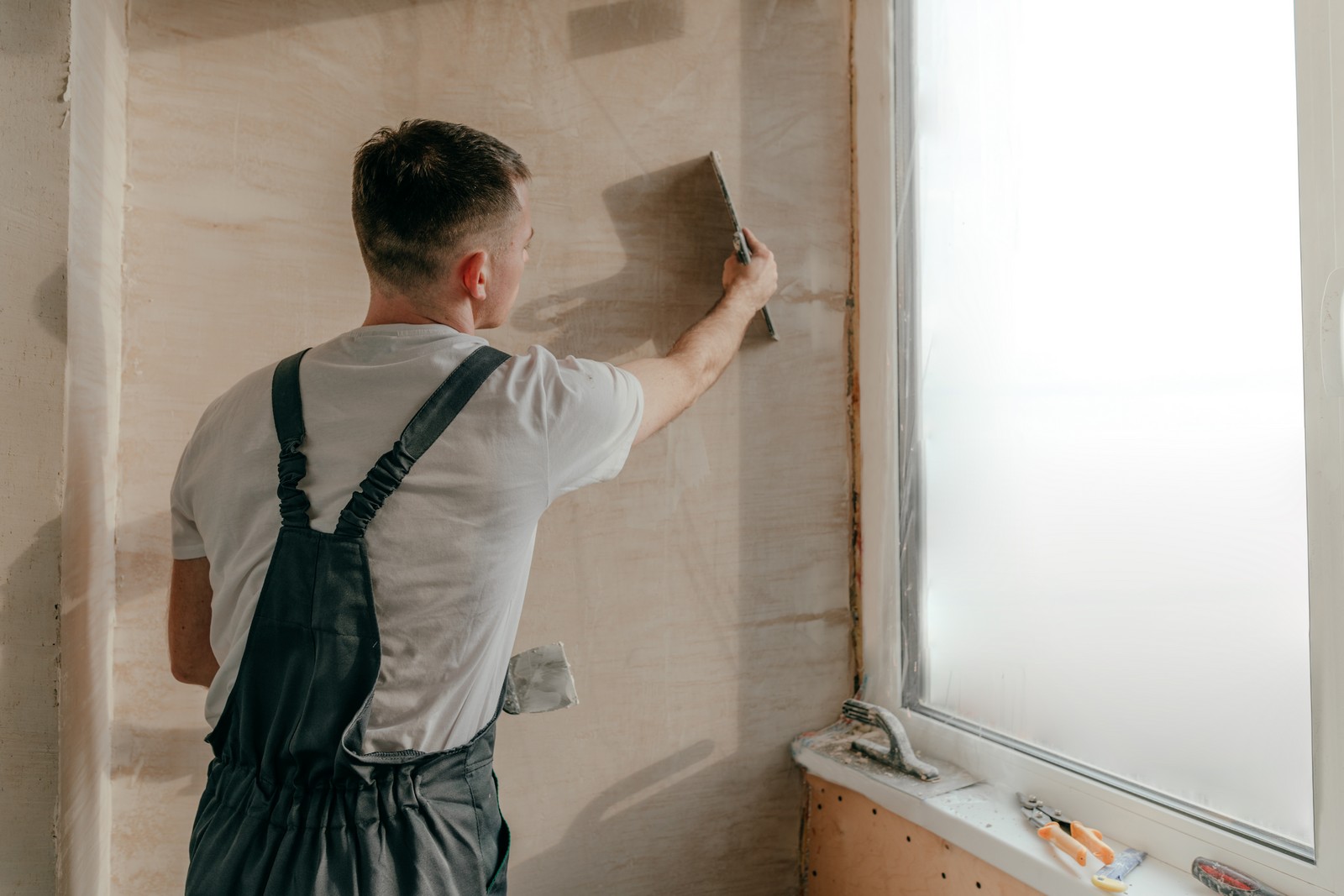 This image shows a person from behind, who appears to be engaged in a home improvement task, specifically preparing a wall for finishing, perhaps for painting or wallpapering. The person is wearing a white t-shirt with dark braces attached to gray work trousers, and is using a spatula or putty knife on the wall. The wall surface looks like it has been recently plastered and is being smoothed out or having imperfections filled. On the windowsill to the right, there are various tools and items related to the task, such as a paint scraper, gloves, and what appears to be a sanding block. The room is bright, with natural light streaming in from the window.