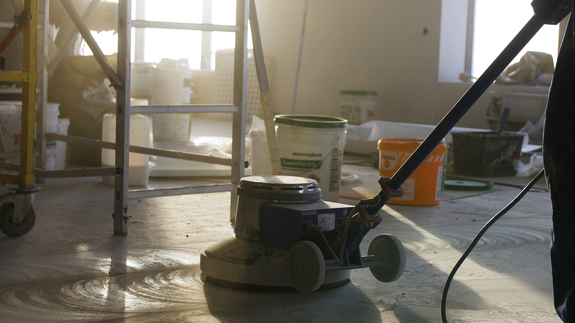 Certainly! Here is a detailed alt text description for the image:
"A construction worker wearing a high-visibility safety vest, hard hat, and protective gloves is kneeling as he polishes a freshly applied mortar floor at a construction site. The worker is using a mechanical floor polisher, which is connected to a nearby power source. The surrounding area is a partially built structure with exposed concrete walls and columns. Building materials, tools, and construction debris are scattered around the site, indicating an active work zone. Natural light filters through the open spaces, casting shadows across the floor and highlighting the worker's focused effort in smoothing the surface."