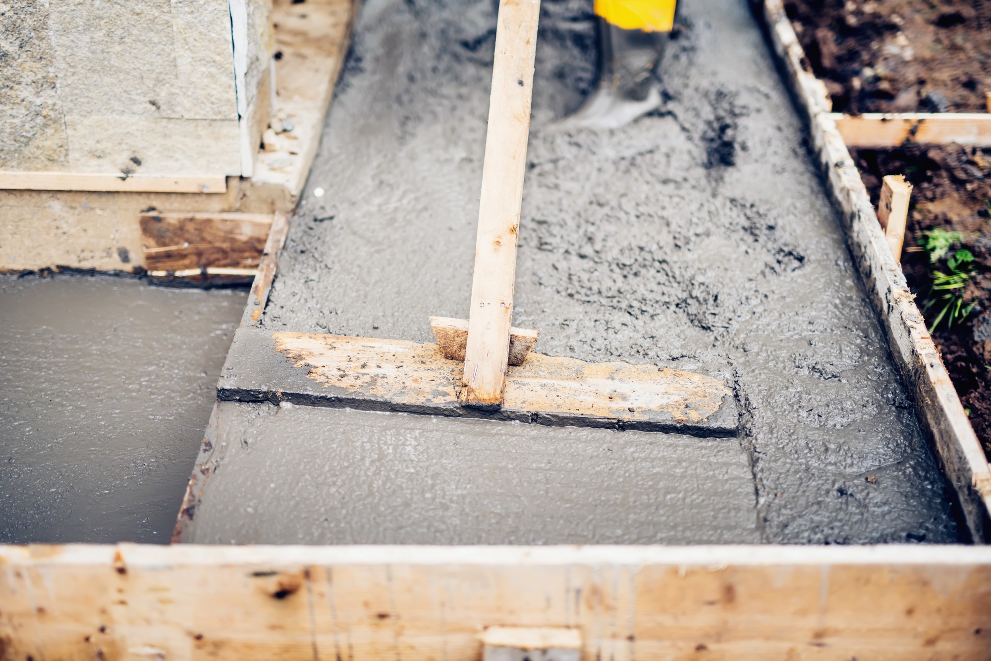 The image shows a skilled mason engaged in the process of building and leveling the first layer of concrete. He is wearing protective gear including a hard hat, safety goggles, and a high-visibility vest. The mason is using a trowel to spread and smooth the freshly poured concrete on what appears to be a foundation or base layer. Surrounding him are various construction tools and a few bags of materials, indicative of a typical building site. In the background, there is a mix of unfinished structures and scaffolding, suggesting an ongoing construction project. The environment is clean and organized, denoting a professional worksite. The mason’s focused expression and precise movements emphasize the level of skill and attention to detail required in this foundational phase of construction. The image highlights the crucial initial steps in creating a strong and level base for future building work.