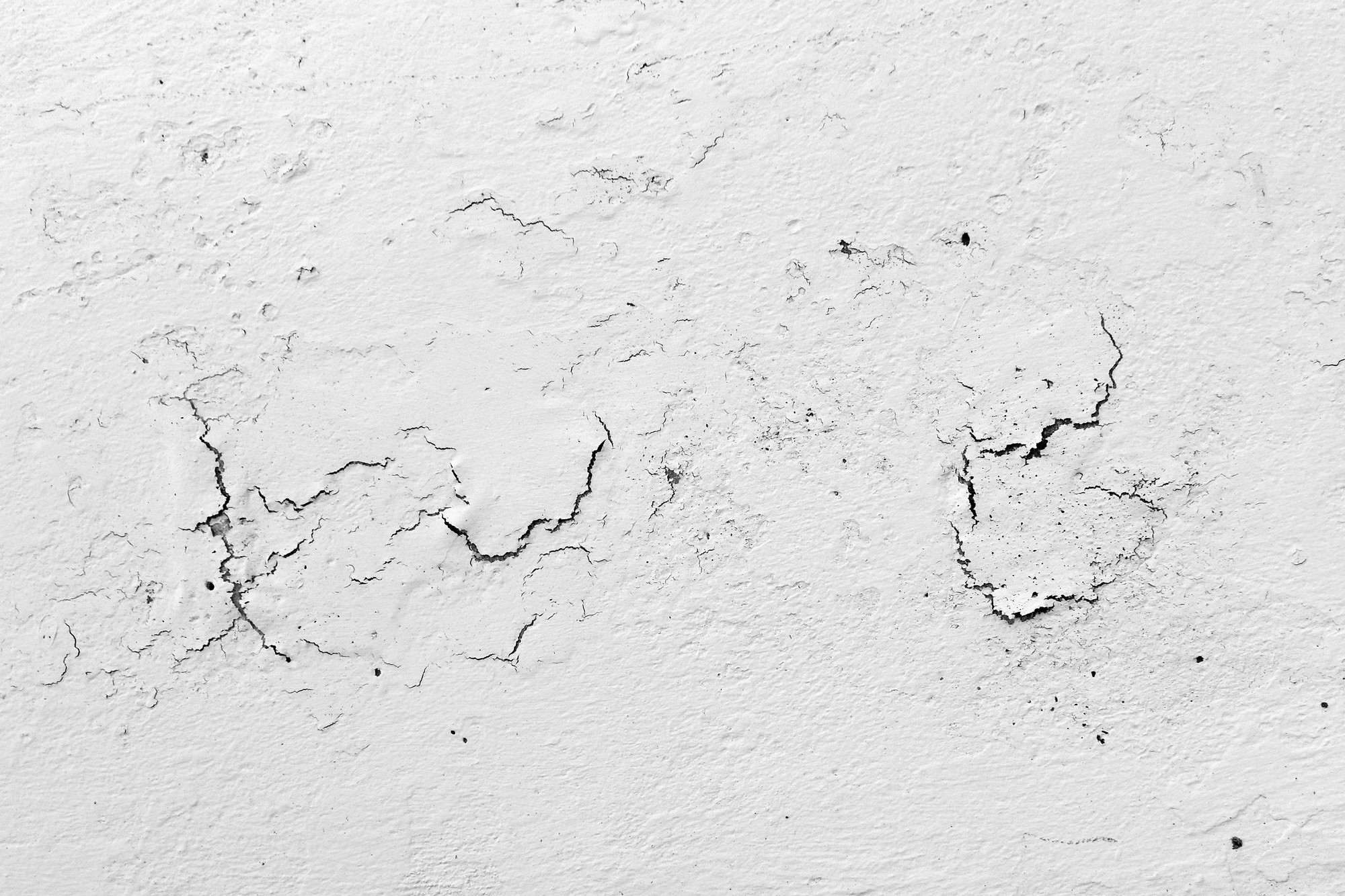 The image shows a close-up view of a deteriorating concrete wall with severe peeling paint. The wall appears to be heavily weathered, with large sections of paint flaking off, revealing the rough, uneven texture of the concrete underneath. The exposed areas are darkened, likely due to prolonged moisture exposure. The presence of water stains and streaks running down the wall indicates ongoing leakage issues, presumably from rain. The overall condition of the wall suggests that it has been subjected to prolonged weathering without adequate maintenance or repair. The visual evidence of water damage and paint degradation underscores the need for urgent remedial action to prevent further deterioration.
